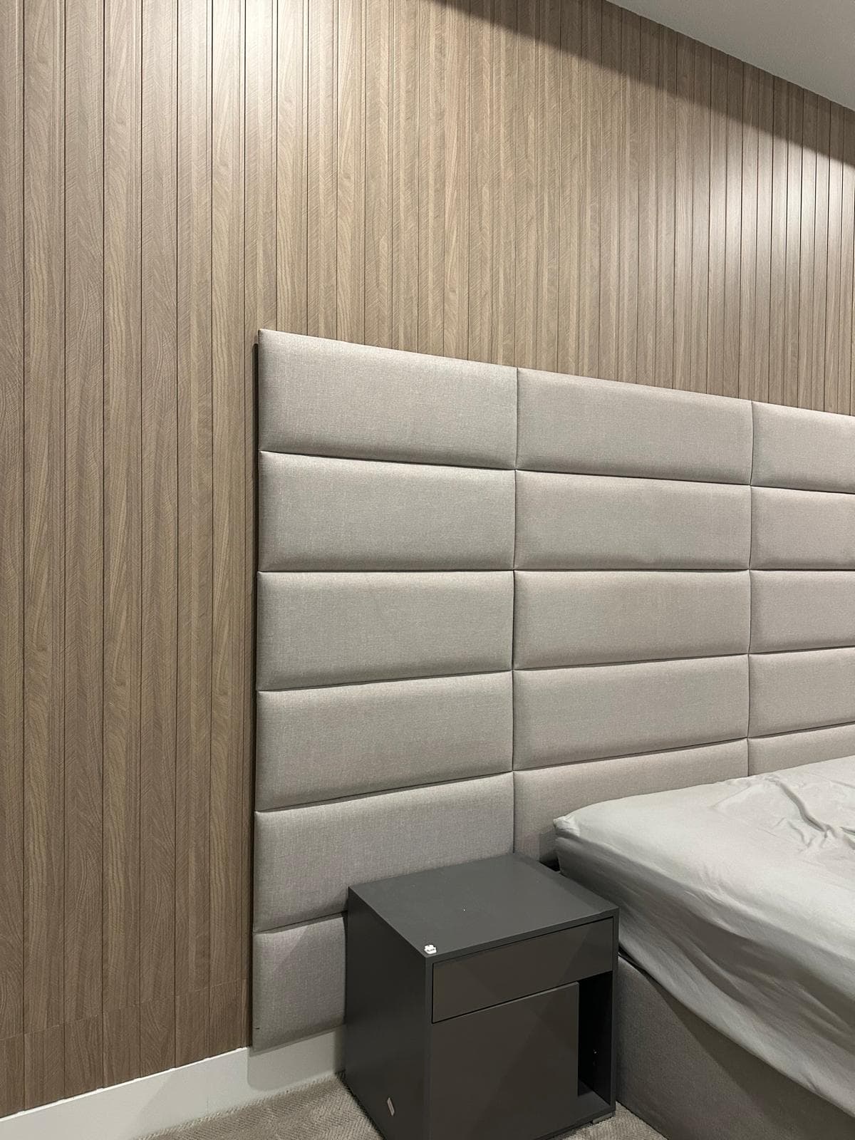 Grey upholstered panel headboard against a vertical wood-slat wall with a dark grey nightstand.