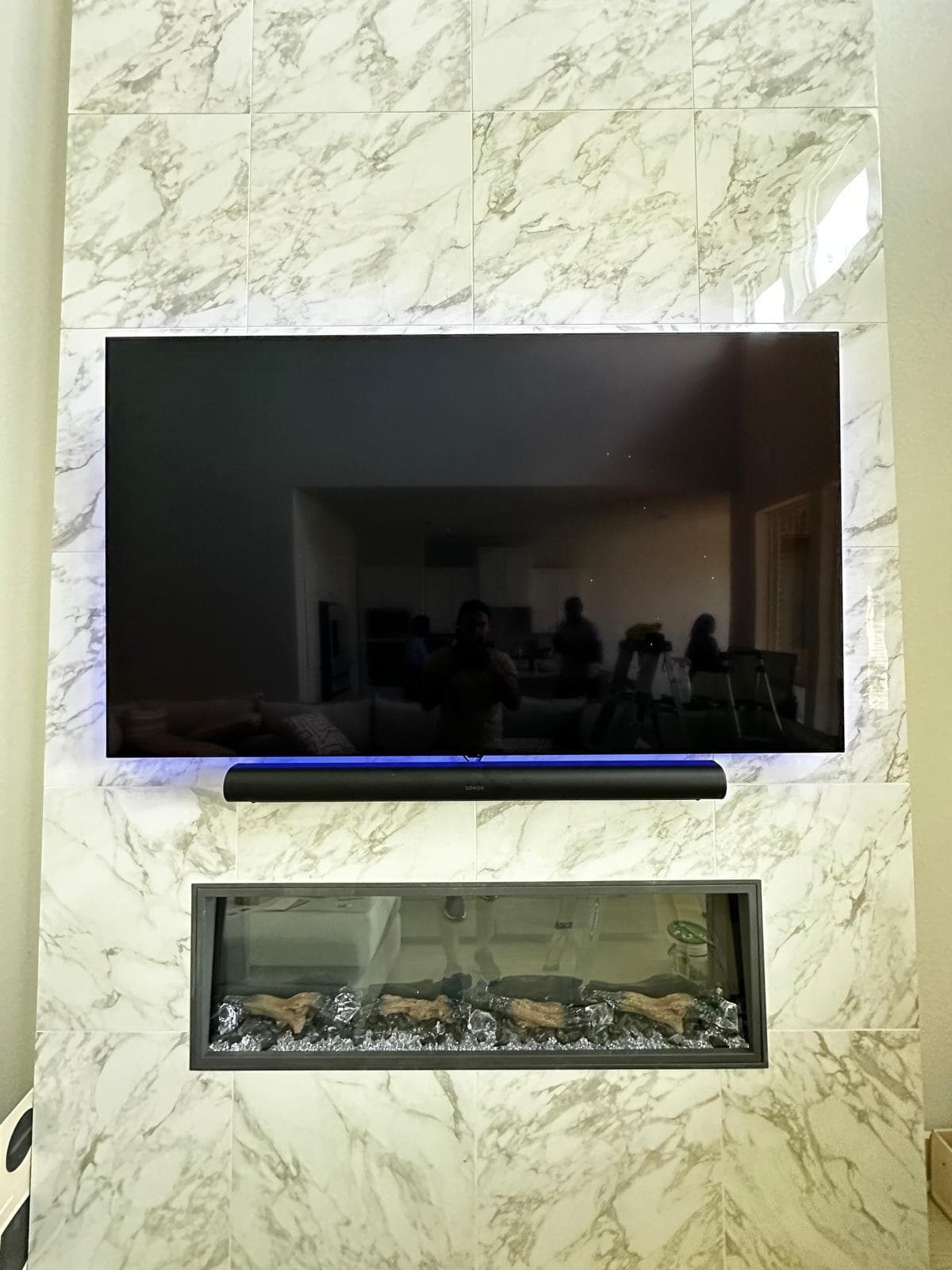 Large TV and soundbar mounted on white marble wall tiles above a modern fireplace.